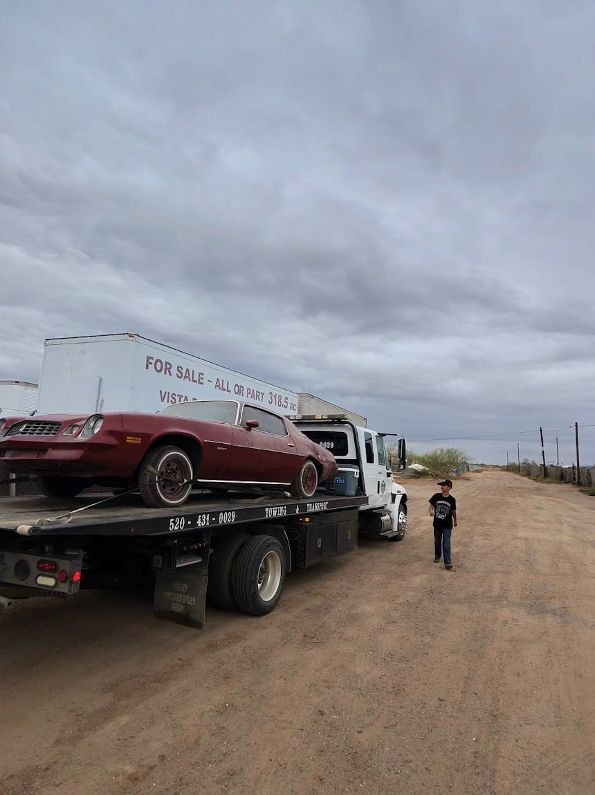 Tow truck carrying a burgundy car on a dirt road with person standing nearby