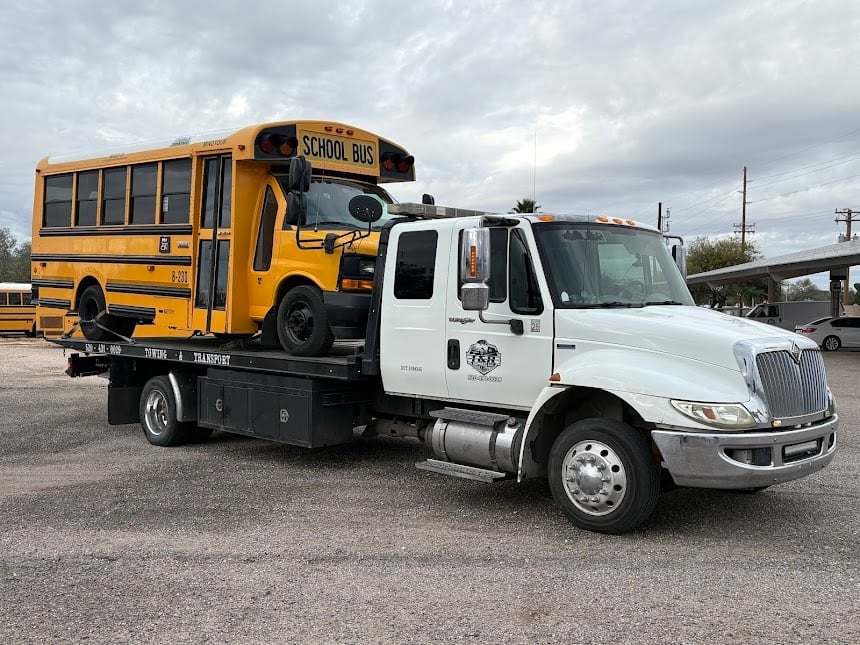 White tow truck carrying a yellow school bus on its flatbed in a parking lot